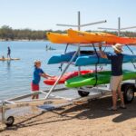 A man and a child loading multiple colourful kayaks onto a multi-level kayak trailer near a lake on a sunny day.
