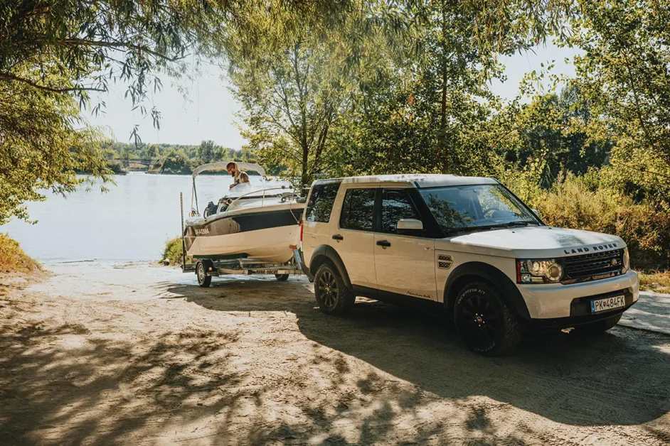 White Land Rover Discovery 4 towing a boat on a trailer towards a lake in a sunny, tree-lined area of New South Wales, Australia, with one people visible on the boat ready for launching.
